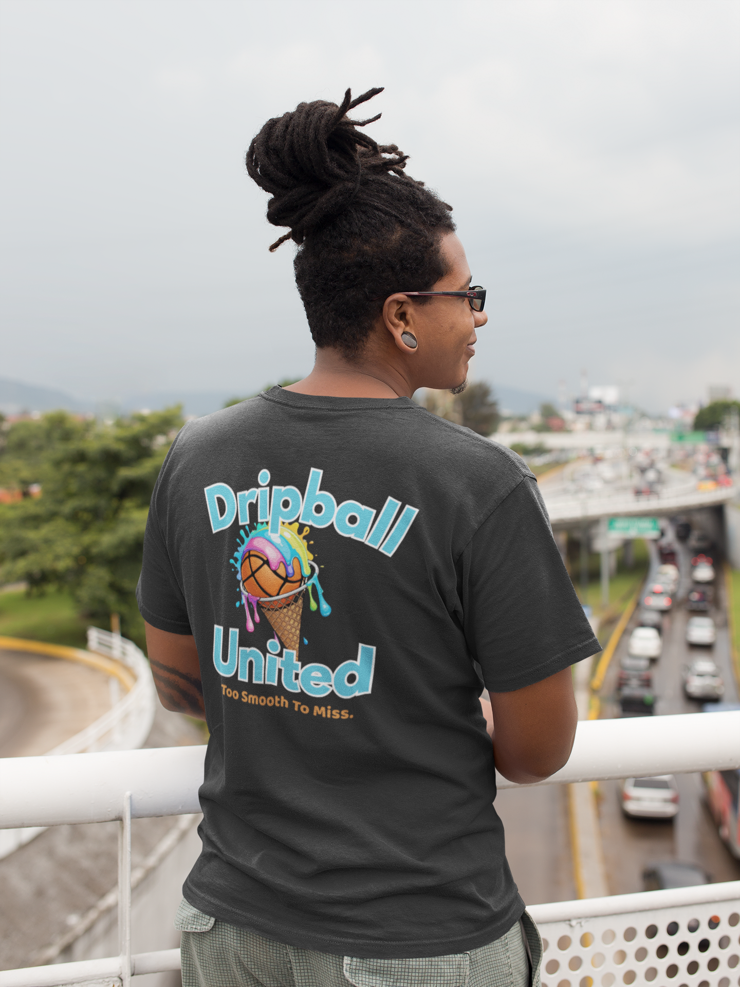 A person with dreadlocks wearing sunglasses and a Dripball United t-shirt stands on an overpass, overlooking a busy street with cars and greenery in the background.