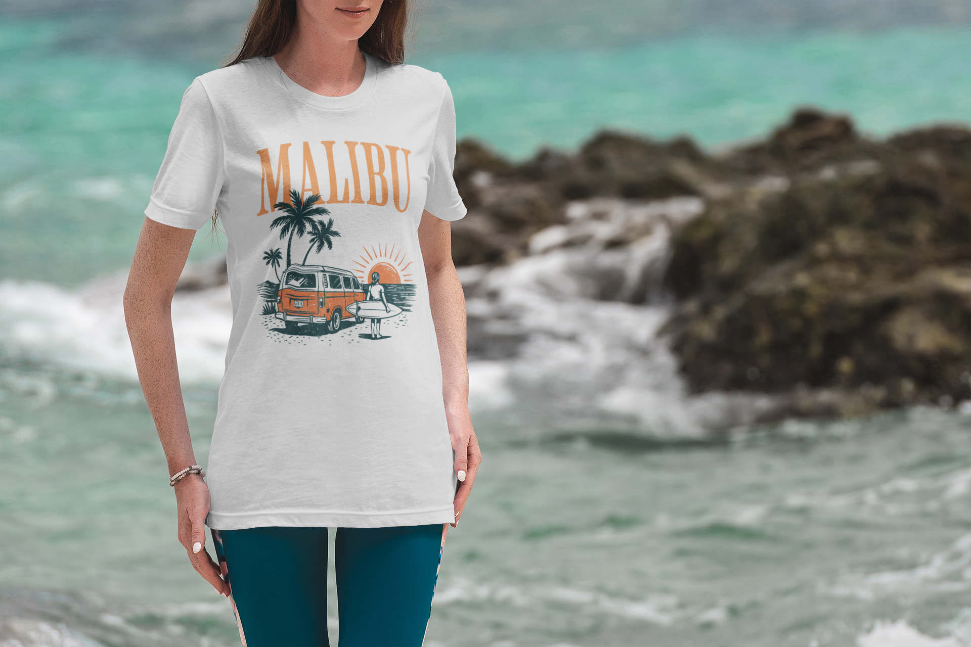 A woman stands by rocky seaside water, wearing a white t-shirt with the word “MALIBU,” a vintage orange van, palm trees, and a surfer graphic printed on it. The ocean and rocks are blurred in the background.